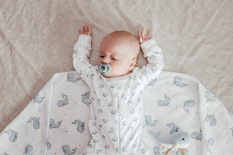 A  baby with a pacifier in their mouth is lying on their back on a cream-colored blanket adorned with a whale pattern.