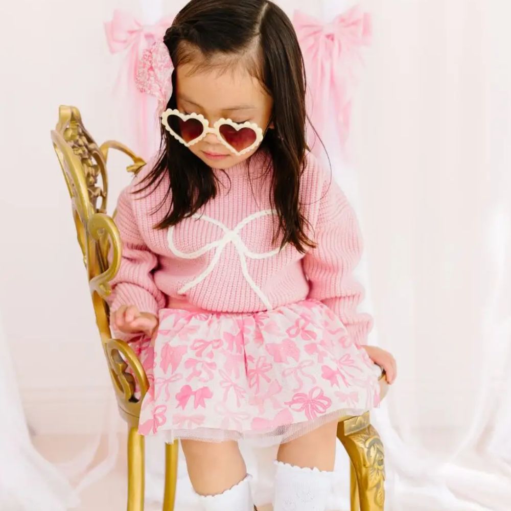 Child wearing a pink outfit with bows, sitting on a gold chair against a white background.