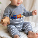 Baby wearing a gray sweater with a train design, holding a wooden toy, on a light-colored surface.