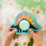 Colorful dinosaur-shaped bath bomb with a green center held by hands against a floral background.