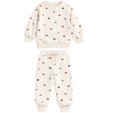 A two-piece children's sweatshirt and jogger pants set in cream, decorated with a scattered pattern of small hearts in muted red, pink, gray, and navy blue. The set features a crewneck sweatshirt with long sleeves and matching elastic-waist jogger pants with drawstrings.