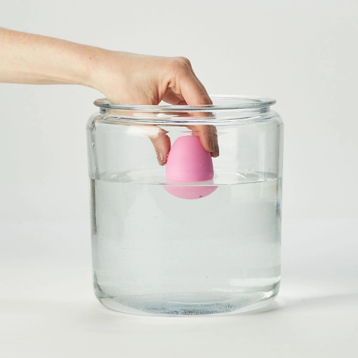 Hand holding a pink egg-shaped object above a glass jar with water on a white background