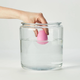 Hand holding a pink egg-shaped object above a glass jar with water on a white background
