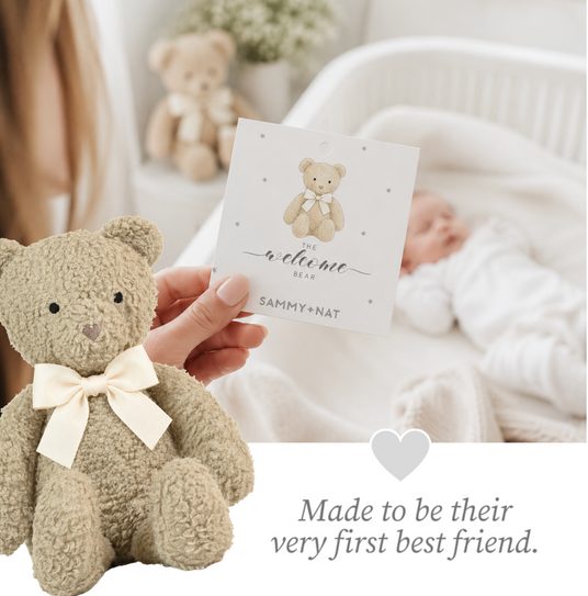 Teddy bear with a bow and a card held by a hand, with a baby in the background.