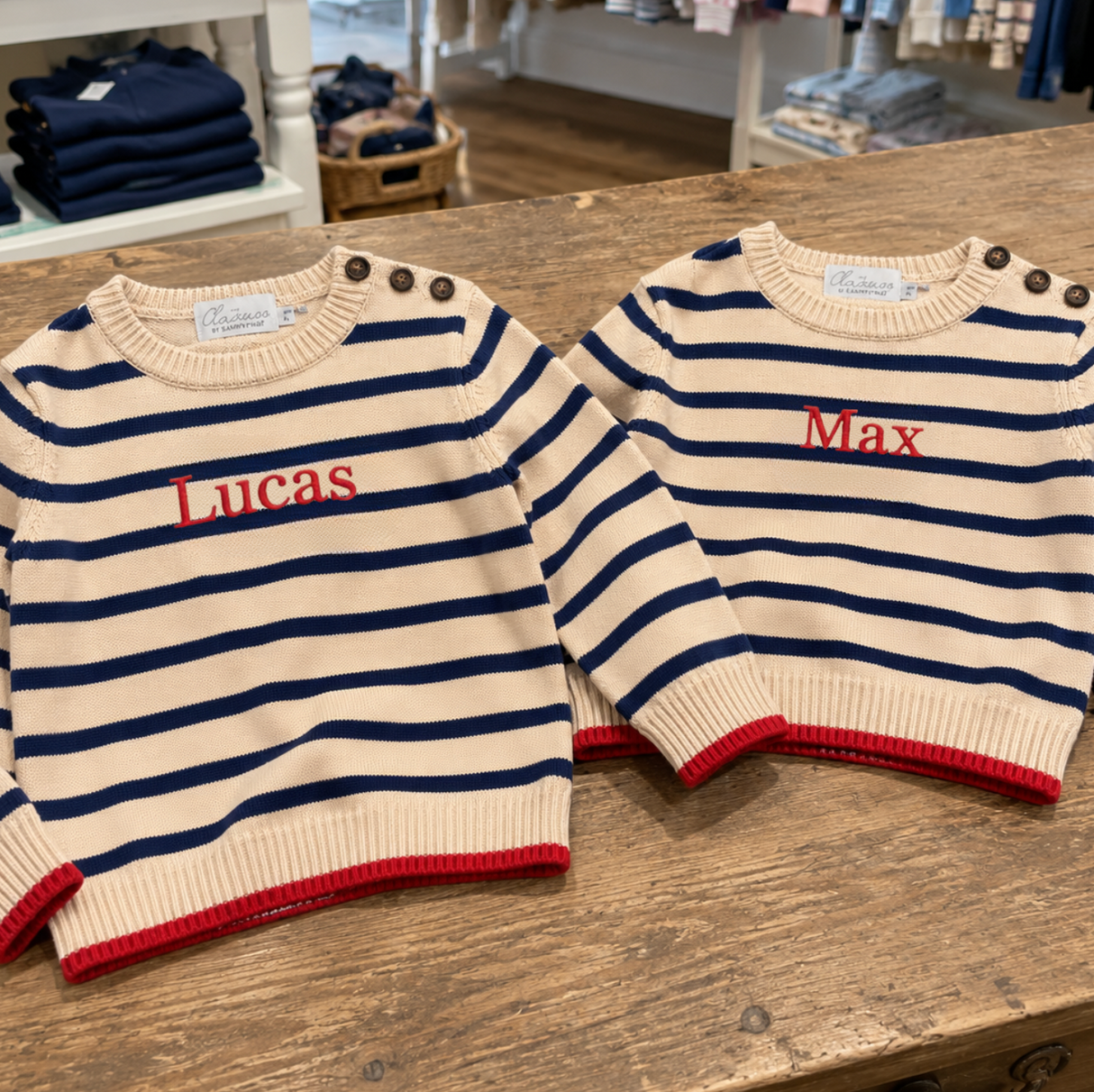 Two striped sweaters with embroidered names on a wooden surface, with a store setting in the background.