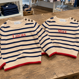 Two striped sweaters with embroidered names on a wooden surface, with a store setting in the background.