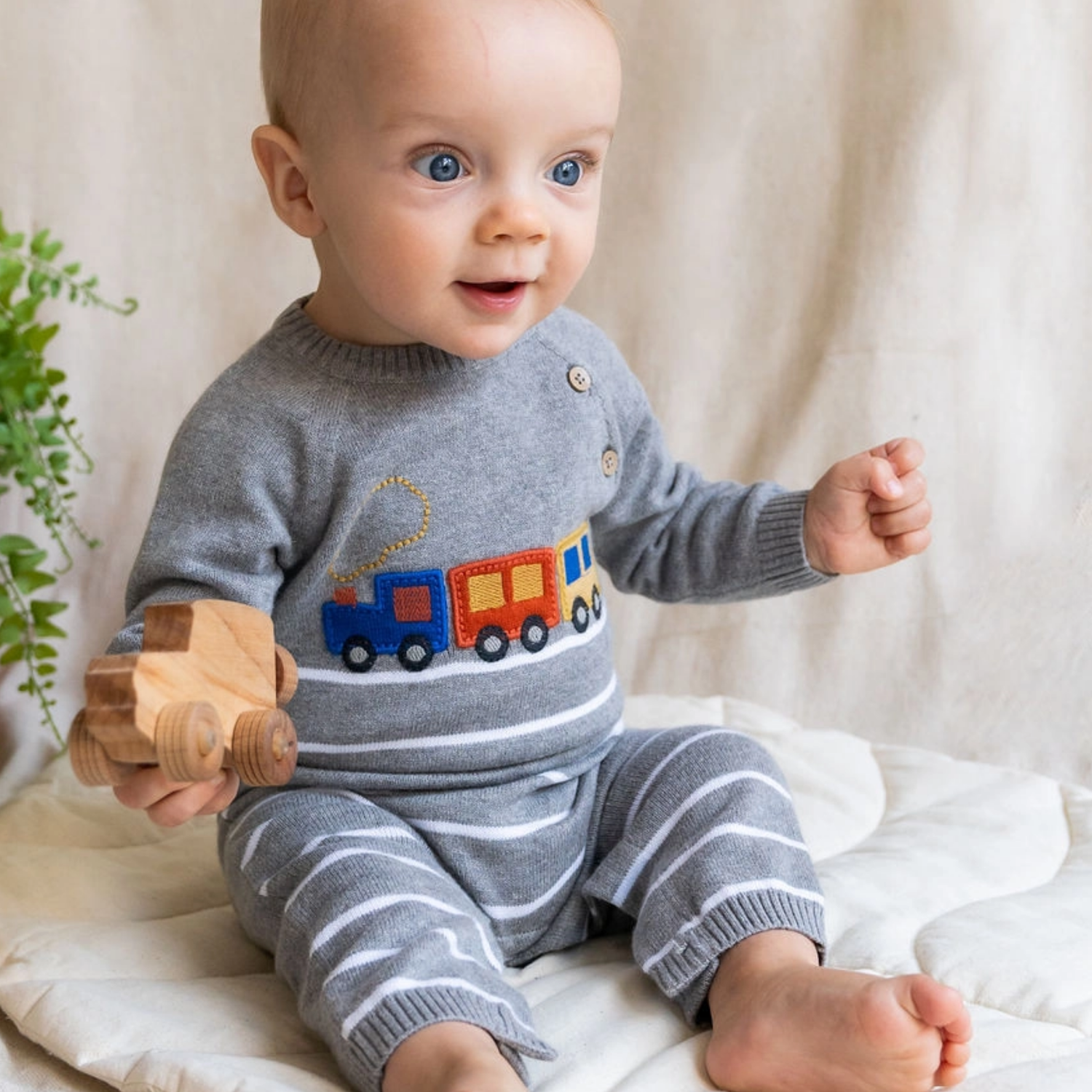 Baby wearing a gray outfit with a train design, holding a wooden toy, on a white blanket.