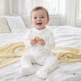 Baby in a white onesie with yellow patterns sitting on a bed.