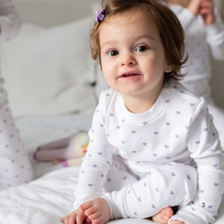 Baby wearing a white onesie with purple heart patterns sitting on a bed.