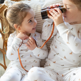 Two children in matching pajamas with a dog pattern, one holding binoculars and the other a leash, sitting on a couch.